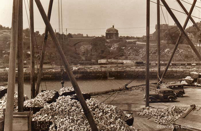 From the top of Granite Pier, we see a lot of going on at Gull Cove. Granite cobblestones, probably from nearby Flat Ledge Quarry, are ready to be loaded onto barges or schooners. Small gauge railroad cars sit on the other side of the cove, ready to haul granite from the quarry on the other side of the Keystone Bridge. Granite Street passes over the bridge, and past the Rockport Granite Company building, which is built with granite, of course!